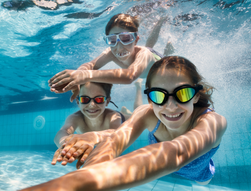 Three kids underwater in an indoor pool, wearing goggles and smiling.