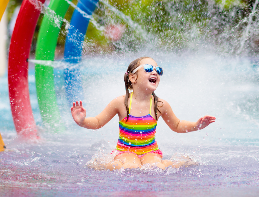 Young girl enjoying a water splash park, wearing a colorful swimsuit and sunglasses.