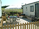 Outdoor patio area with a table, chairs, and an umbrella next to a static caravan in a British holiday park, enclosed by a wooden fence with a view of grassy fields in the background.